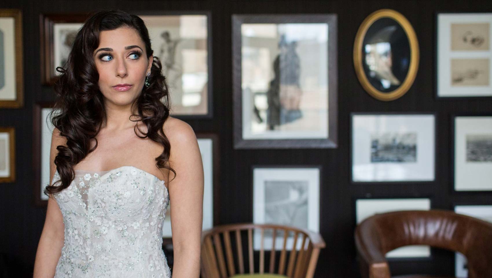 Natalie looking off to the side in her wedding dress in front of a wall of framed photos and portraits