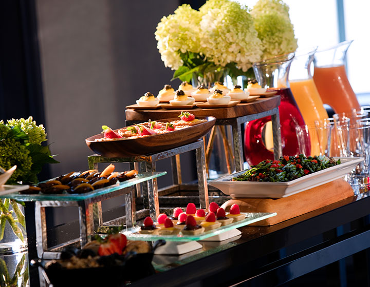 Banquet food placed on tiered trays with floral pieces next to mugs of juice